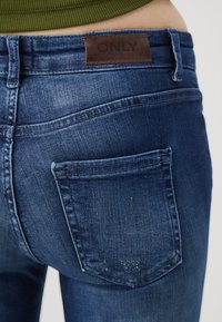 Mørkeblå denimjeans med et brunt lærmerke, to baklommer og en lett falmet tekstur. Synlig sømdetalj langs sømmene.