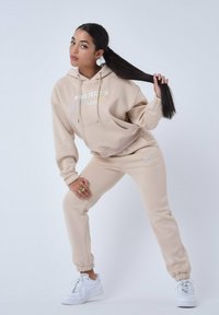 Beige hoodie met een voorzak en kleurrijk logo, gecombineerd met bijpassende taps toelopende broek; heeft elastische manchetten en is gestyled met witte sneakers.