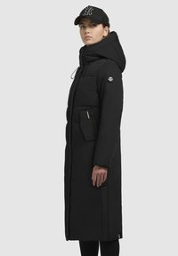 Žena u dugom crnom kaputu s kapuljačom i crnom bejzbol kapom, stoji u profilu ispred jednostavne pozadine.