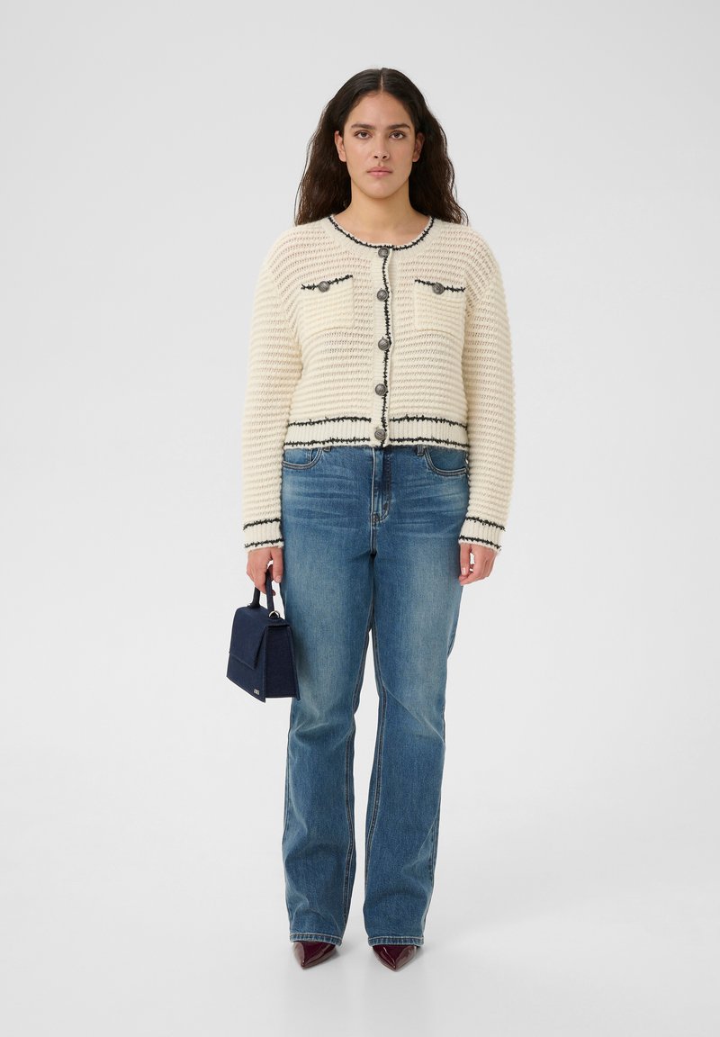 Strickjacke in Creme mit schwarzen Akzenten, runder Ausschnitt und Knopfverschluss; kombiniert mit ausgestellten blauen Jeans und kleiner navy Handtasche.