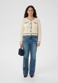 Kvinna som bär en krämfärgad stickad kofta med svarta kanter och stora knappar, blå jeans, vinröda spetsiga skor, håller en liten marinblå handväska.