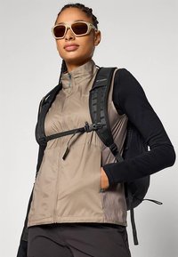 Ελαφρύ μπεζ γιλέκο με φερμουάρ και ρυθμιζόμενα λουράκια, σε συνδυασμό με ένα μαύρο μακρυμάνικο μπλουζάκι. Το μοντέλο φορά γυαλιά ηλίου και μια μαύρη τσάντα πλάτης.