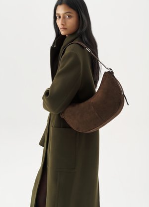 Borsa a spalla in suede marrone con design curvo e sottile tracolla, indossata sopra un cappotto verde scuro con grandi tasche frontali.