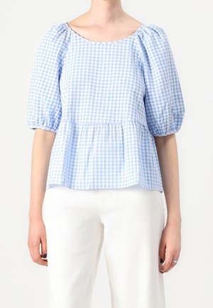 Kobieta w jasno niebieską i białą kraciastą bluzkę z bufkami na rękawach i białe spodnie na jednolitym tle.
