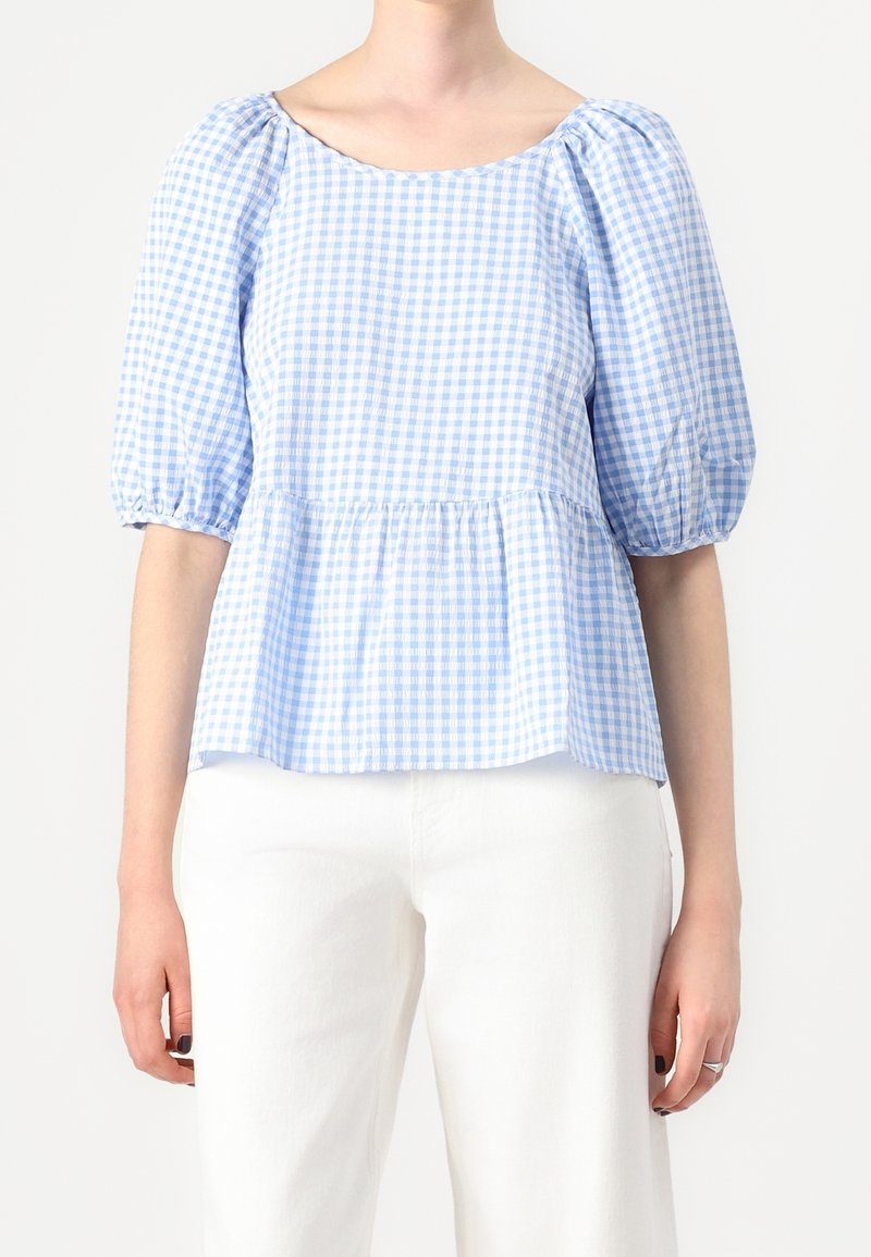 Femme portant un chemisier à carreaux bleu clair et blanc avec des manches bouffantes et un pantalon blanc sur un fond uni.