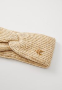 Knit headband in light beige with a ribbed texture and a knotted design. Features a small gold logo accent.