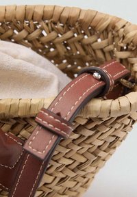 Panier en osier tressé avec un revêtement en tissu beige clair, doté d'une poignée en cuir marron avec des coutures blanches et d'un accent en métal argenté.