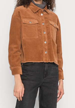 Kvinde iført en cropped brun fløjlsjakke med sølvknapper over en grå rullekrave og sorte jeans.
