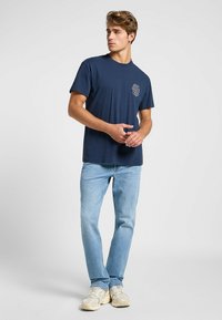 Camiseta de algodón azul marino con mangas cortas y cuello redondo; presenta un pequeño diseño de logo blanco en el lado izquierdo del pecho; combinada con jeans de color azul claro.