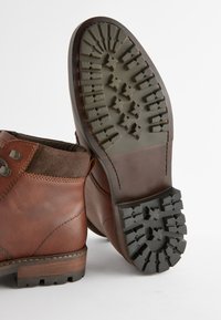 Hnedé kožené členkové topánky s textúrovaným semišovým golierom, okrúhlym špičkou a gumovou podrážkou s výrazným vzorom protišmykov.