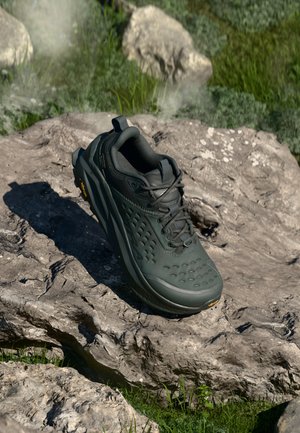 Sapatilha de caminhada verde escuro com parte superior texturizada, protuberâncias proeminentes na sola e biqueira arredondada. Design leve com calcanhar reforçado e fecho com atacadores.