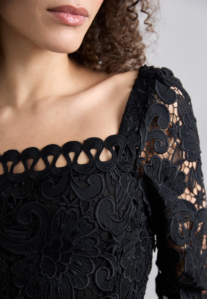 Czarna bluzka z koronką, zdobiona kwiatowym haftem, z kwadratowym dekoltem wykończonym dekoracyjną lamówką oraz długimi rękawami z misternymi wzorami.