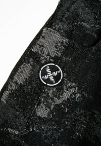 Textil negro con un patrón texturizado, primer plano de un parche de tela contrastante con un logotipo redondo en blanco y negro y detalles de costura.