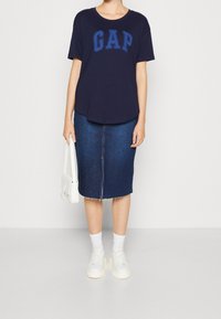 Námořnické tričko s logem "GAP", spárované s tmavými denimovými midi sukněmi. Bílé tenisky s robustními podešvemi a malou bílou kabelkou.