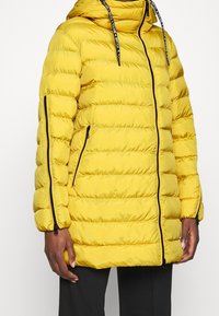 Chaqueta acolchada amarilla con costuras horizontales, capucha, bolsillos con cremallera y ribete de cremallera negro. Presenta puños elásticos y capucha con cordón ajustable.