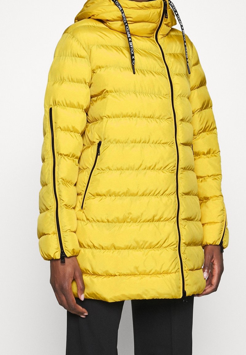 Chaqueta acolchada amarilla con costuras horizontales, capucha, bolsillos con cremallera y ribete de cremallera negro. Presenta puños elásticos y capucha con cordón ajustable.