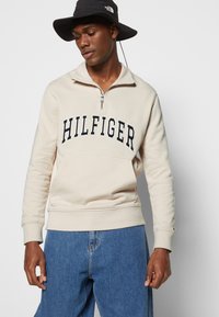 Felpa con zip color crema con il logo "HILFIGER" sul petto, dotata di tasca a marsupio e polsini a coste, abbinata a jeans azzurri chiari.