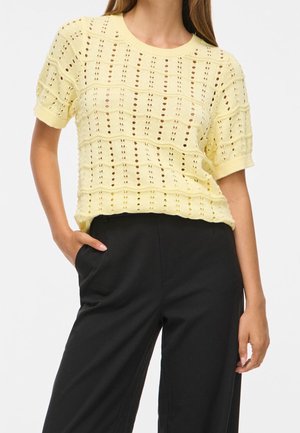 Femme portant un top en maille jaune clair à manches courtes avec un motif ajouré et un pantalon noir taille haute, une main dans la poche.