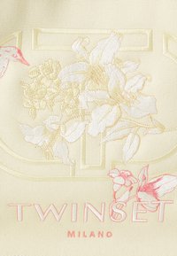 Il tessuto crema presenta un intricato ricamo floreale in tonalità pastello. Il logo "TWINSET MILANO" è in evidenza in un rosa chiaro.