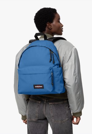 Blauwe stoffen rugzak met een voorvak met rits, verstelbare schouderbanden en een Eastpak logo patch. Draagt over een grijze bomberjack.