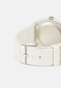 Montre en silicone blanche avec un bracelet texturé et une boucle en argent. Elle présente un boîtier rond en métal avec un fond transparent et un logo gravé.