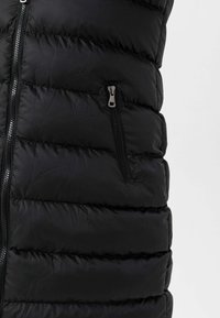 Sort puffer vest med kviltet overflade, der har en sidelomme med lynlås og glat tekstur. Lynlåsen lukker vertikalt foran.