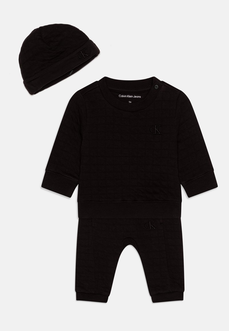 Črno bombažno dvodelno oblačilo z dolgem rokavom, ki ima pritrdilne gumbe in elastiko na manšetah, v kombinaciji z usklajenimi hlačami in kapico.