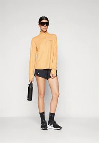 Šviesiai oranžinis ilgomis rankovėmis užsegamas viršus, juodos trumpikės, juodi sportbačiai ir tamsūs akiniai nuo saulės. Laiko juodą gertuvę. Paprasto dizaino, sportiško stiliaus.