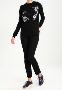 Pull noir avec des motifs floraux blancs et le logo "Guess", associé à un pantalon noir ajusté et des chaussures bleu marine. Tissu lisse et coupe ajustée.