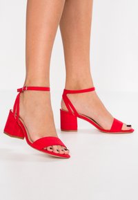 Sandales à talons blocs en daim rouge avec sangle autour de la cheville, bout ouvert et détail en évidement circulaire sur les côtés. Présente une finition lisse et texturée.