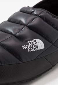 Black synthetic slip-on shoe featuring a quilted texture, padded upper, and a prominent white logo. The sole is textured for grip.
