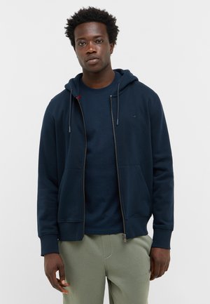 Navy Zip-Hoodie mit einer Vordertasche, verstellbaren Zugbändern und gerippten Bündchen, kombiniert mit einem dunklen Shirt und hellgrünen Hosen.