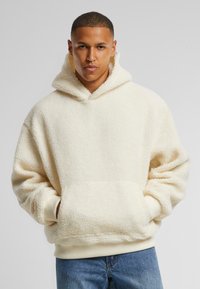 Cremefarbener Fleece-Hoodie mit großer Fronttasche, gerippten Bündchen und oversized Schnitt, ausgestattet mit einem hohen Ausschnitt und strukturiertem Oberflächen.