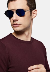 Para czarnych okularów przeciwsłonecznych w lekko niebieskich szkłach, o owalnym kształcie, noszona z burgundowym swetrem z okrągłym dekoltem. Gładka tekstura, minimalistyczny design.
