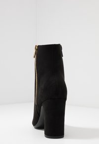 Bota de tobillo de ante negro con un alto tacón cuadrado y una cremallera dorada lateral. Textura suave y diseño de punta redondeada.