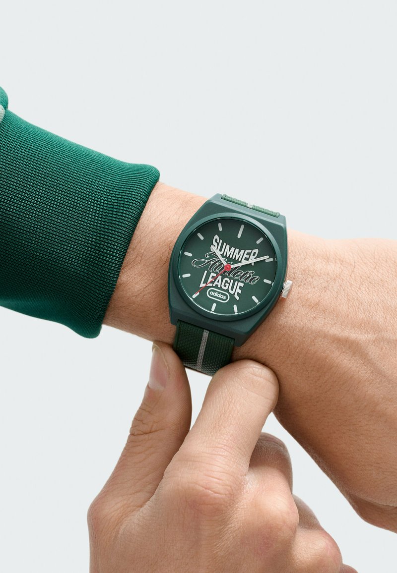 Persona ajustando un reloj de pulsera Adidas verde que muestra "Liga Atlética de Verano" en la esfera con marcadores blancos y aguja de segundos roja.