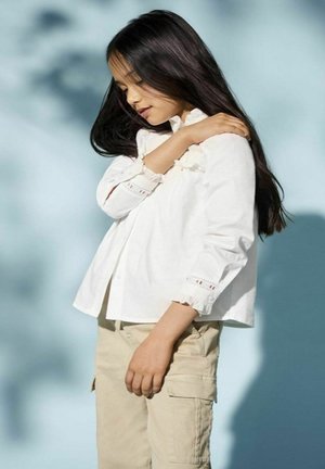 Witte blouse met gerimpelde manchetten en kantdetails, gecombineerd met beige cargobroeken, te zien tegen een zachte blauwe achtergrond.