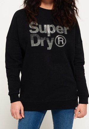 Person trägt einen schwarzen Sweatshirt mit silbernem "Super Dry®"-Schriftzug, kombiniert mit blauen Jeans, steht vor weißem Hintergrund.