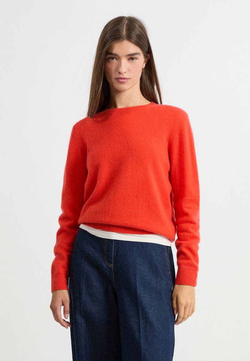 Pull en laine rouge avec un col rond et des manches longues, associé à un pantalon en denim bleu foncé taille haute. Tissu doux et texturé.
