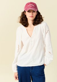Witte blouse met lange mouwen en een V-hals, versierd met borduursels op de mouwen, gecombineerd met een stofroze baseballpet met de tekst "CALIFORNIA."