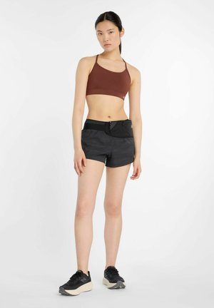 Ung kvinde står iført brun sports-bh, sorte atletikshorts med bæltetaske og sorte løbesko mod hvid baggrund.