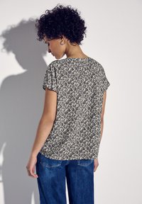 Donna con capelli ricci che indossa una maglietta a maniche corte con un motivo bianco e nero e jeans blu, in piedi con le spalle rivolte verso un muro bianco.