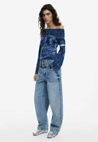 Blaues schulterfreies Sweatshirt mit strukturiertem Muster, kombiniert mit übergroßen, hellblauen Jeans. Das Model trägt silberne Sneakers.