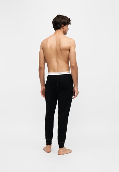 Homme debout pieds nus de dos, torse nu, portant un pantalon de jogging noir avec une ceinture élastique blanche sur un fond clair uni.