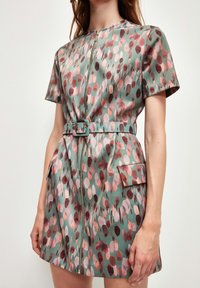 Robe verte à manches courtes avec une fermeture éclair à l'avant et une taille ceinturée, ornée d'un motif de pois roses et bordeaux, agrémentée de poches latérales.