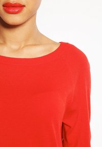 Kvinna iklädd en ljus röd tröja och matchande röd läppstift, visar hals och del av axeln mot en vit bakgrund.