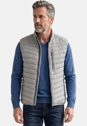 Mann mit grauem Haar und Bart, der eine hellgraue gesteppte Weste über einem blauen Langarmpullover und dunklen Jeans trägt und zur Seite schaut.
