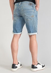 Shorts en denim bleu clair avec des revers, cinq poches et un patch en cuir marron à la taille. Portés avec des baskets noires et blanches.