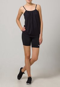 Kvinde iført en sort ærmeløs top med spaghetti-stropper, sorte Nike shorts og sorte atletiske sko, stående med det ene ben krydset over det andet.