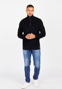 Schwarzer Reißverschluss-Pullover mit gerippter Textur, der einen hohen Kragen und flache Nähte aufweist, kombiniert mit hellblauen Jeans und weißen Sneakern.